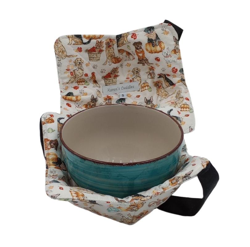 Bowl cozies are hot pads designed to hold hot or cold bowls, has a versatile black strap for hand support and easy storage. Doting cute dogs in a fall setting of leave and pumpkins on a soft cream fabric. Made of 100% cotton fabric, ideal for reheating food in the microwave and doubling as a placemat. This cozy is available in small, holding soup bowls up to 6.5” diameter or medium pasta bowls holding up to 7.5” diameter.